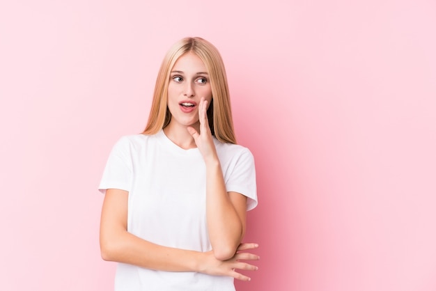 Joven rubia sobre fondo rosa está diciendo una noticia secreta de frenado en caliente y mirando a un lado