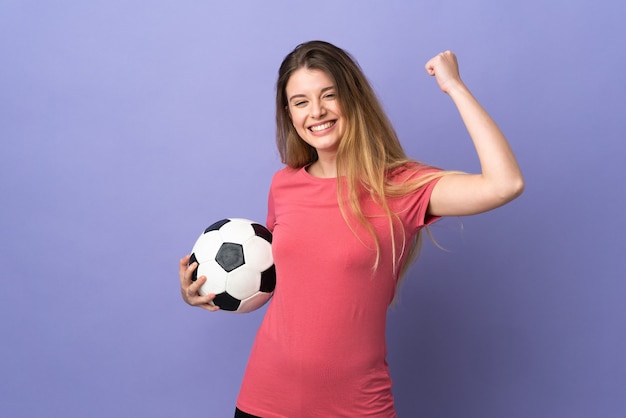 Joven rubia sobre pared aislada con balón de fútbol celebrando una victoria