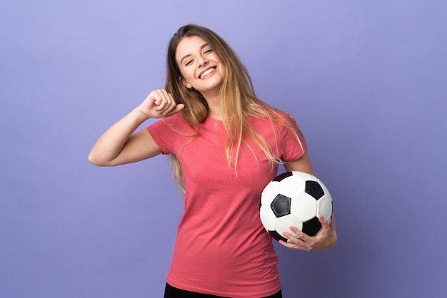 Joven rubia sobre pared aislada con balón de fútbol y orgulloso de sí mismo