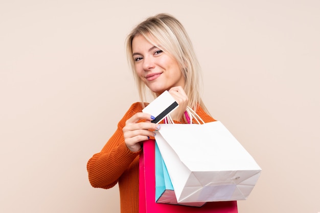 Joven rubia sobre pared aislada sosteniendo bolsas de compra y una tarjeta de crédito