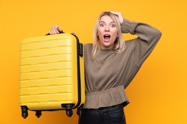 Joven rubia sobre pared amarilla aislada en vacaciones con maleta de viaje y sorprendida