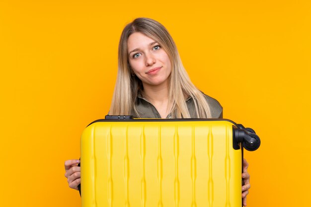 Joven rubia sobre pared amarilla aislada en vacaciones con maleta de viaje