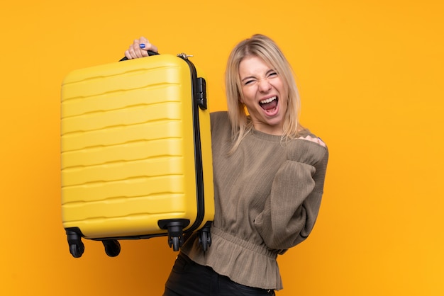 Joven rubia sobre pared amarilla aislada en vacaciones con maleta de viaje