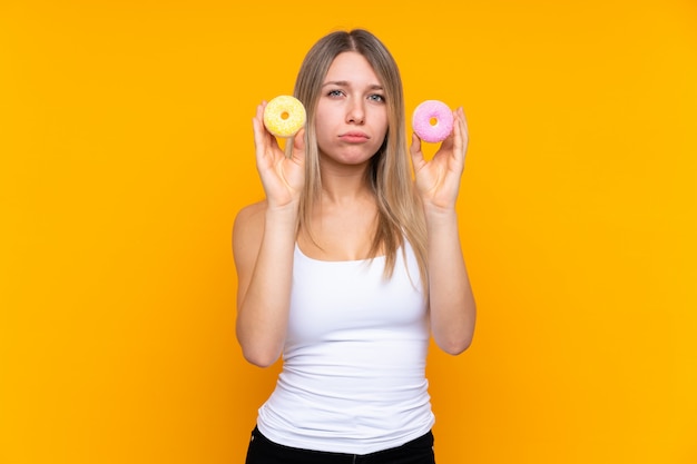 Joven rubia sobre pared azul aislada con donas con expresión triste