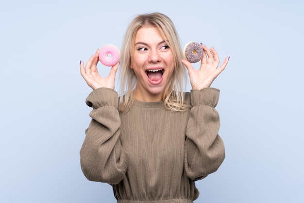 Joven rubia sobre pared azul aislada con donas y sorprendido