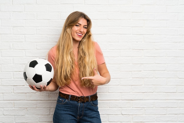 Joven rubia sobre pared de ladrillo blanco sosteniendo un balón de fútbol