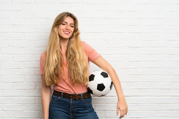 Joven rubia sobre pared de ladrillo blanco sosteniendo un balón de fútbol