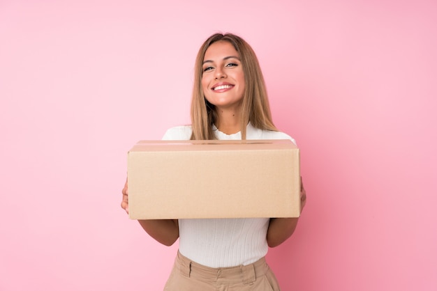 Joven rubia sobre pared rosa aislada sosteniendo una caja para moverla a otro sitio