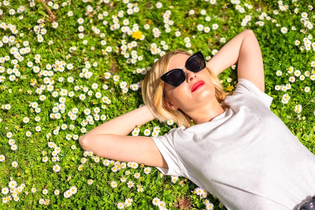 Una joven rubia con sombrero y gafas de sol respirando pelo puro en primavera en un parque en la naturaleza de la ciudad tumbada en el césped junto a las margaritas parte del espacio de copia