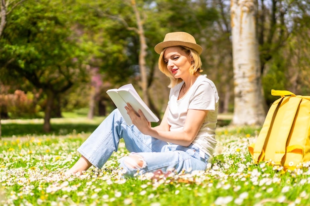 Una joven rubia con un sombrero leyendo un libro en primavera en un parque de vacaciones en la ciudad junto a la naturaleza y junto a las margaritas sentadas en el césped