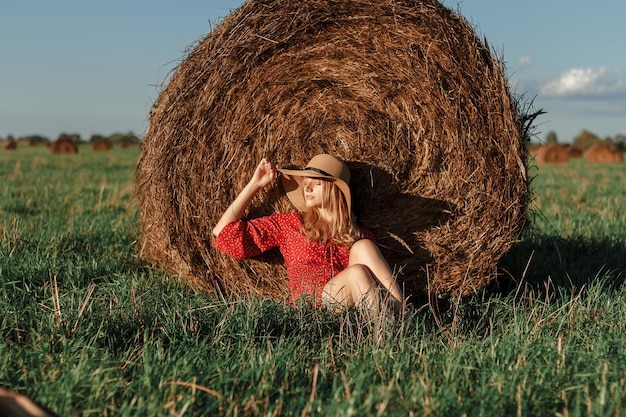 Una joven rubia con un sombrero de paja se sienta junto a un pajar en un momento de calma atmosférica