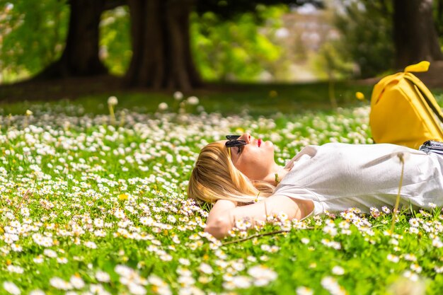 Una joven rubia con un sombrero que se relaja y respira cabello puro en la primavera en un parque en la naturaleza de la ciudad tirada en el césped junto a las margaritas mirando la cámara