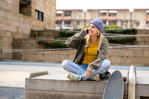 Joven rubia con sombrero veri peri coolor hablando con un amigo usando un teléfono inteligente adolescente patinadora sentada en un banco en el espacio de copia de la ciudad