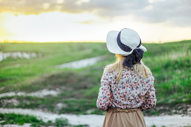 Una joven rubia con un sombrero y un vestido histórico entre las colinas se encuentra en el camino.