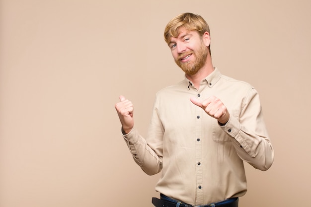 Joven rubia sonriendo alegremente y casualmente apuntando hacia un lado, sintiéndose feliz y satisfecho por encima de la pared