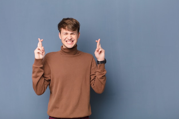 Joven rubia sonriendo y cruzando ansiosamente ambos dedos, sintiéndose preocupado y deseando o esperando buena suerte aislado contra la pared plana