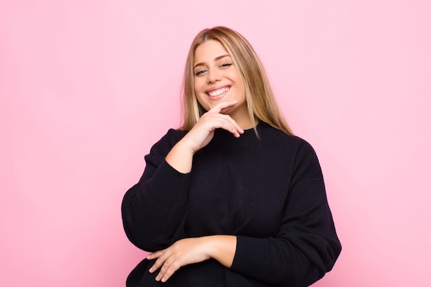 Joven rubia sonriendo, disfrutando de la vida, sintiéndose feliz, amigable, satisfecha y despreocupada con la mano en la barbilla contra la pared plana