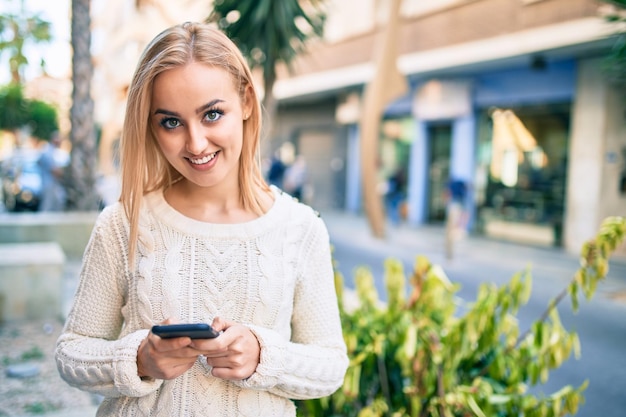 Joven rubia sonriendo feliz usando un teléfono inteligente en la ciudad