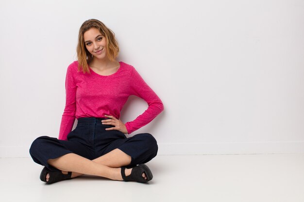 Joven rubia sonriendo felizmente con una mano en la cadera y una actitud segura, positiva, orgullosa y amigable contra la pared de color plano