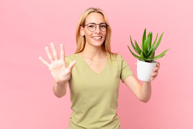 Joven rubia sonriendo y luciendo amigable, mostrando el número cinco y sosteniendo un cactus