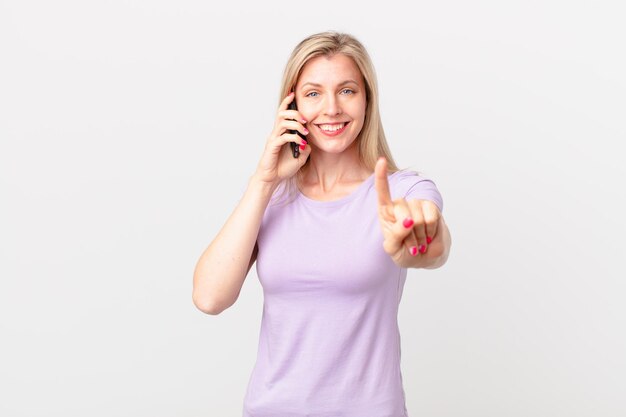 Joven rubia sonriendo con orgullo y confianza haciendo el número uno y llamando con un teléfono inteligente