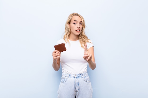 Joven rubia sonriendo con orgullo y confianza haciendo la pose número uno triunfante, sintiéndose como un líder con una billetera