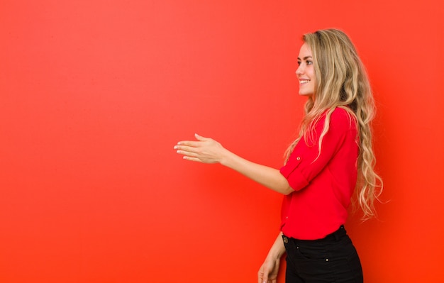 Joven rubia sonriendo, saludando y ofreciendo un apretón de manos para cerrar un acuerdo exitoso, concepto de cooperación en la pared roja