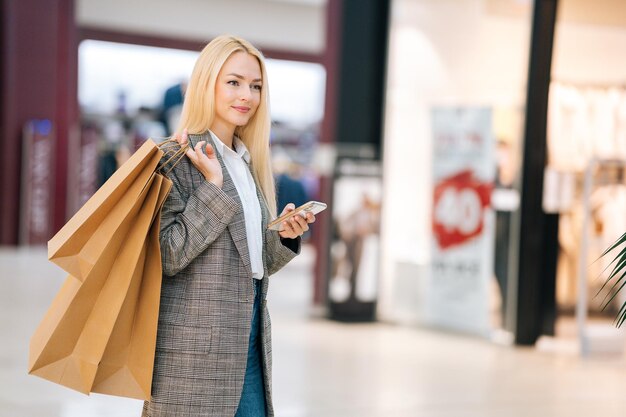 Una joven rubia sonriente y atractiva con un abrigo elegante sosteniendo un teléfono móvil y bolsas de papel de compras con compras mirando hacia otro lado en el centro comercial. Bonito mensaje de escritura femenino en el teléfono inteligente en la tienda.