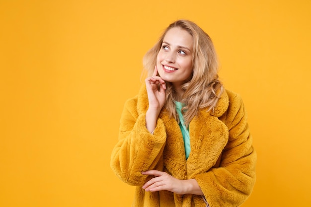 Una joven rubia sonriente y pensativa con un abrigo de piel amarillo posando aislada en un retrato de estudio de fondo de pared naranja. Concepto de estilo de vida de las emociones de las personas. Simulacros de espacio de copia. Coloque la mano apoyada en la barbilla.