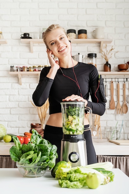 Joven rubia sonriente en ropa deportiva negra escuchando música y haciendo batido verde en la cocina de casa