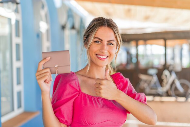 Foto joven rubia sosteniendo una billetera al aire libre con los pulgares hacia arriba porque algo bueno sucedió