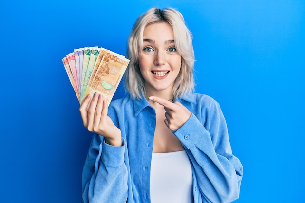 Joven rubia sosteniendo billetes en pesos filipinos sonriendo feliz señalando con la mano y el dedo