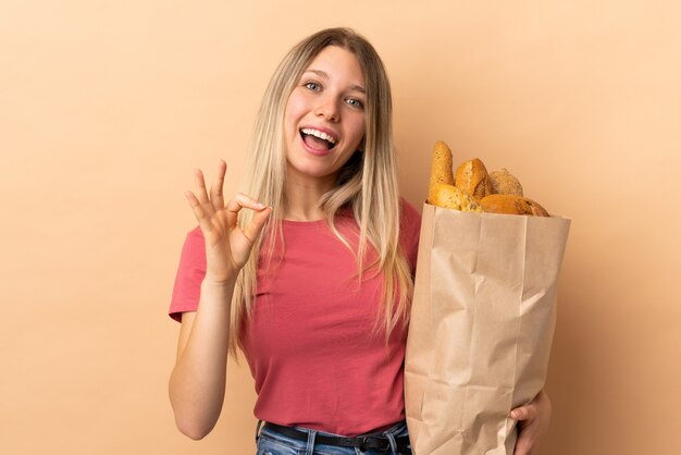 Joven rubia sosteniendo una bolsa llena de panes aislados en la pared de color beige que muestra un signo bien con los dedos