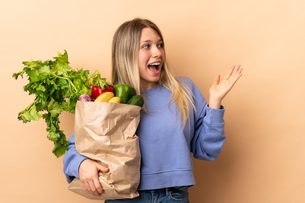 Joven rubia sosteniendo una bolsa llena de verduras sobre pared aislada con expresión facial sorpresa