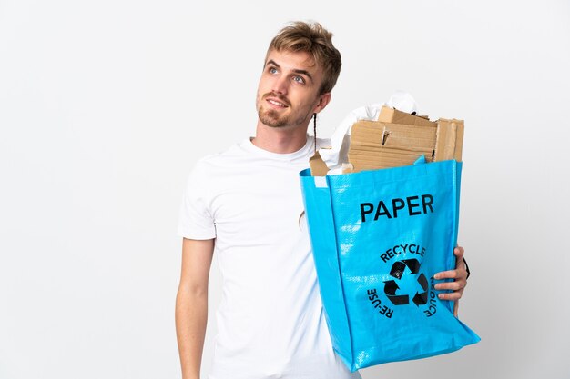 Joven rubia sosteniendo una bolsa de reciclaje llena de papel para reciclar aislado en blanco pensando en una idea mientras mira hacia arriba