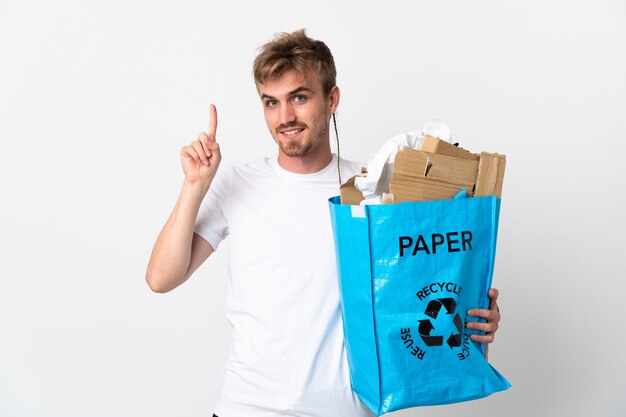 Joven rubia sosteniendo una bolsa de reciclaje llena de papel para reciclar aislado sobre fondo blanco apuntando hacia una gran idea