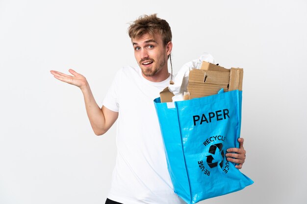 Joven rubia sosteniendo una bolsa de reciclaje llena de papel para reciclar aislado sobre fondo blanco con expresión facial sorprendida