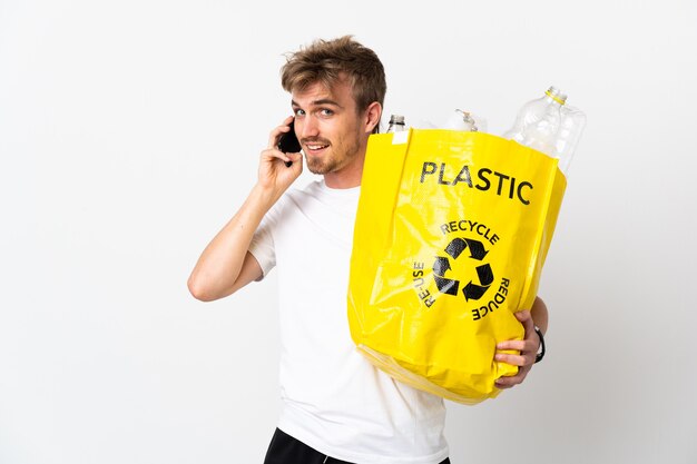 Joven rubia sosteniendo una bolsa de reciclaje llena de papel para reciclar aislado sobre fondo blanco manteniendo una conversación con el teléfono móvil con alguien