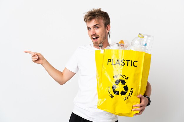 Joven rubia sosteniendo una bolsa de reciclaje llena de papel para reciclar aislado sobre fondo blanco sorprendido y apuntando con el dedo hacia el lado