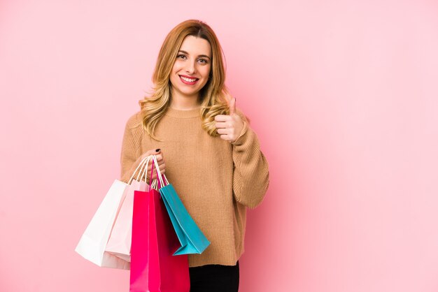 Joven rubia sosteniendo bolsas de compras sonriendo y levantando el pulgar