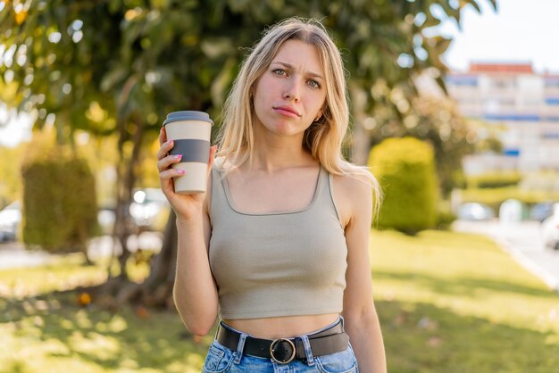 Joven rubia sosteniendo un café para llevar al aire libre con expresión triste