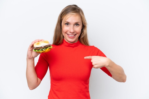 Joven rubia sosteniendo una hamburguesa aislada de fondo blanco con expresión facial sorpresa