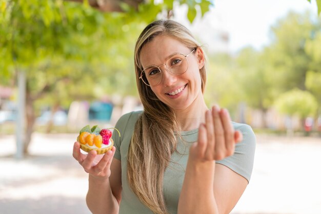 Foto joven rubia sosteniendo una tartaleta al aire libre invitando a venir con la mano feliz de que hayas venido