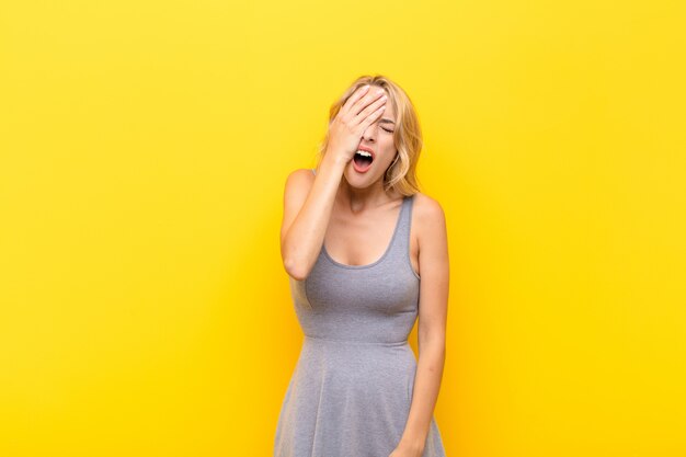 Joven rubia con sueño, aburrida y bostezando, con dolor de cabeza y una mano cubriendo la mitad de la cara contra la pared naranja