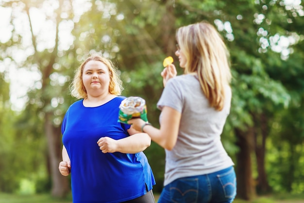 Una joven rubia de tallas grandes corre en el parque cerca de una mujer comiendo patatas fritas