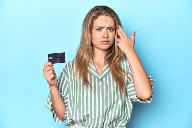 Joven rubia con una tarjeta de crédito en un estudio azul mostrando un gesto de decepción con el dedo índice