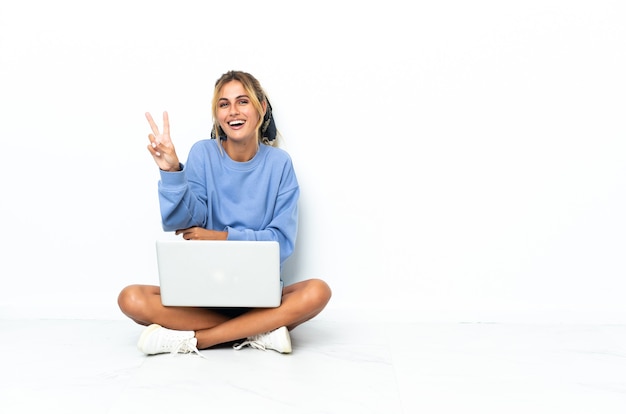 Joven rubia uruguaya con el portátil aislado en la pared blanca sonriendo y mostrando el signo de la victoria