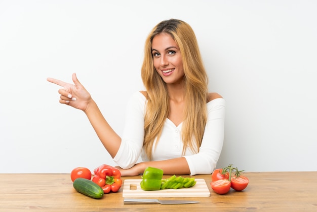 Joven rubia con verduras en una mesa apuntando con el dedo al lado