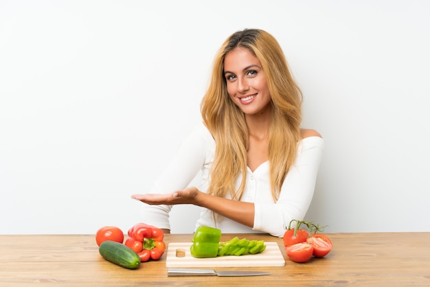 Joven rubia con verduras en una mesa extendiendo las manos a un lado para invitar a venir