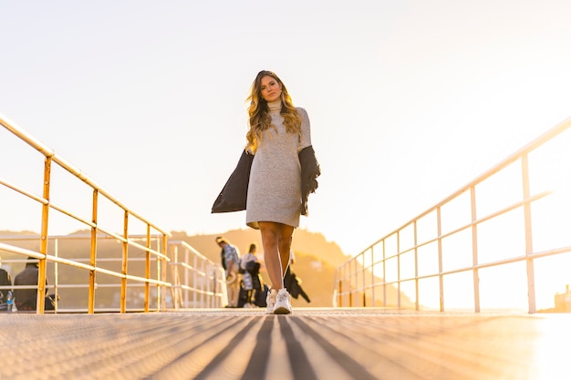 Una joven rubia vestida de gris en un puerto en una hermosa puesta de sol naranja